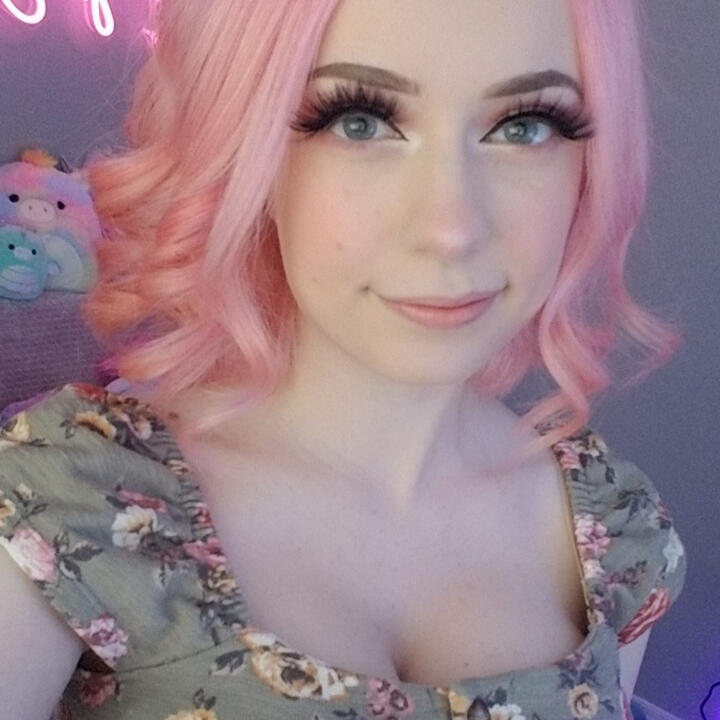 A circle-cropped image of SquishShe. She has short pink hair that's been curled. She is wearing a light green shirt with some white and pink roses here and there. Closed mouth smile looking at you.